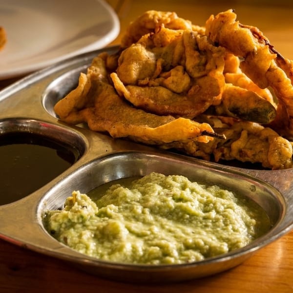 Crispy fried pakoras and chili pakoras served with dark and light green chutneys on a metal plate.