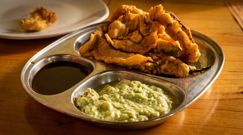 Crispy fried pakoras and chili pakoras served with dark and light green chutneys on a metal plate.