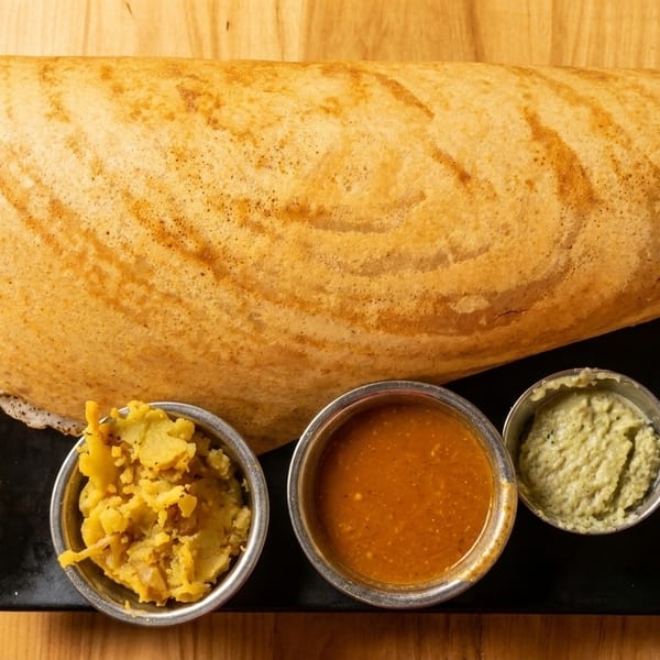 A golden-brown dosa with three small bowls of chutney and potato curry on a black plate.